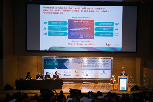 Simpósio Internacional, registro de uma convidada enquanto ministra uma palestra com um telão no fundo.
